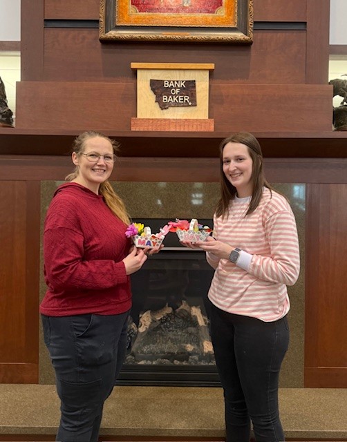 Two Bank of Baker employees pose in front of the bank's fireplace and logo with handcrafted May Day baskets that they made for the local nursing home residents